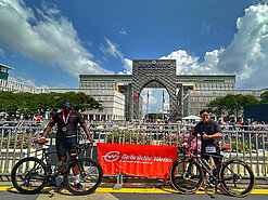 Two Gebrüder Weiss employees in Malaysia pose outdoors with their bikes in front of an orange Gebrüder Weiss banner. There's a large architectural arch in the background. 