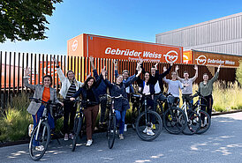 Gebrüder Weiss employees outdoors with their bikes pose with their arms up to celebrate the end of the cycling campaign. There are two Gebrüder Weiss orange truck trailers in the background. 