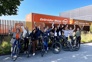 Gebrüder Weiss employees outdoors with their bikes pose with their arms up to celebrate the end of the cycling campaign. There are two Gebrüder Weiss orange truck trailers in the background. 