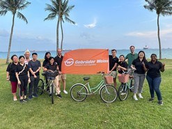 Gebrüder Weiss Employees in Singapore stand outside in front of a Gebrüder Weiss sign with a bike. Palm trees and the ocean are in the background. 