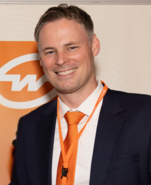 Smiling businessman with short dark hair, orange tie, white shirt and black jacket standing in front of a Gebrüder Weiss logo