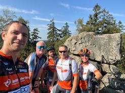 Gebrüder Weiss Employees in the Czech Republic outdoors in their cycling gear. There's a boulder, pine trees and a blue sky in the background. 